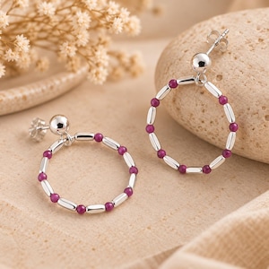 Pode incluir: Um par de brincos de prata com design de argola circular. Cada argola é composta por pequenas contas magenta alternadas com espaçadores cilíndricos de prata lisa, suspensos por um pino de esfera de prata polida com fecho de borboleta.