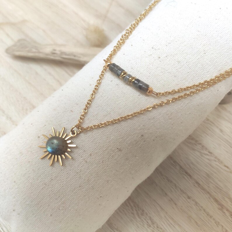 May include: A double-layered gold chain necklace featuring a sun-shaped pendant with a central iridescent labradorite stone. The upper chain is adorned with a series of small, dark grey rectangular beads separated by tiny gold-toned metal spacers.