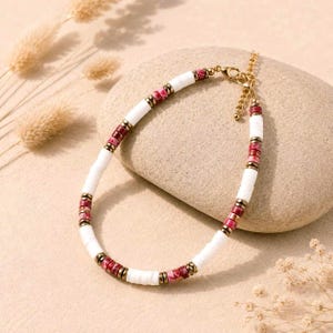 Op de afbeelding: Een armband met afwisselende segmenten van gladde witte cilindrische kralen en kleinere, getextureerde rood-roze stenen schijfjes. De kralen worden gescheiden door dunne, goudkleurige metalen tussenstukjes en het sieraad heeft een goudkleurige karabijnsluiting.