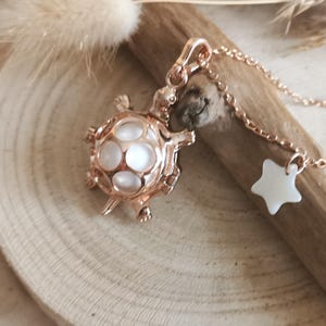 May include: Rose gold-colored turtle pendant necklace with a white star charm. The turtle pendant features a cage-like design with white, pearl-like inserts. The necklace is displayed on a wooden surface, showcasing the jewelry's intricate details.