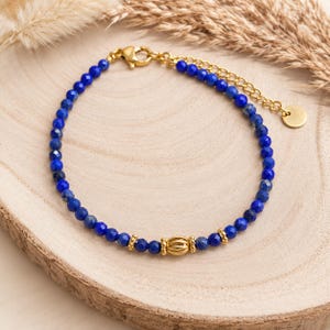 Peut inclure: Un bracelet délicat composé de petites perles facettées en lapis-lazuli bleu profond montées sur une chaîne dorée. Le bijou présente une perle centrale dorée striée, deux petites entretoises dorées, un fermoir mousqueton et une chaînette.