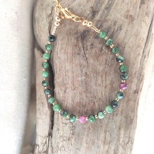 May include: A delicate gold-toned bracelet featuring a strand of green and black gemstone beads interspersed with small gold beads. Two pink gemstone beads add a pop of color. The bracelet is secured with a gold clasp.