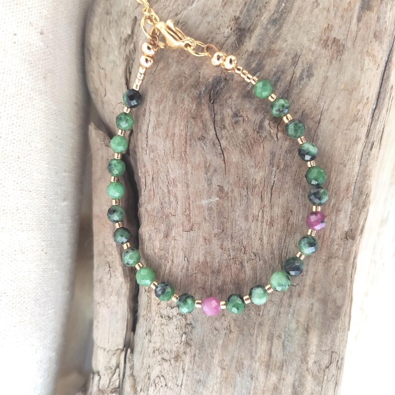 May include: A delicate gold-toned bracelet featuring a strand of green and black gemstone beads interspersed with small gold beads. Two pink gemstone beads add a pop of color. The bracelet is secured with a gold clasp.