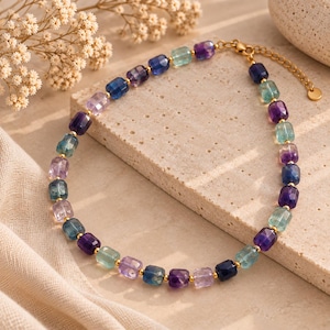 Pode incluir: Um colar de contas com pedras preciosas de fluorite facetadas em forma de barril em tons de roxo, azul e verde. As contas são separadas por pequenos espaçadores de metal dourado e o colar inclui uma corrente de metal dourado com fecho.
