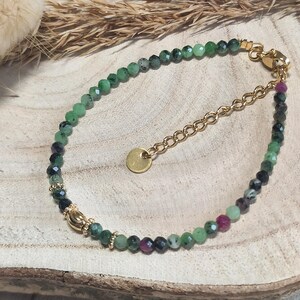 Op de afbeelding: Een delicate armband met kleine, gefacetteerde kralen in groen en robijnrood. De kralen zijn geregen op een goudkleurige ketting en sluiting. Een kleine, ronde gouden bedel hangt aan de ketting.