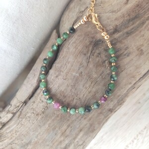 May include: A delicate bracelet featuring green and dark green beads interspersed with small pink beads. The beads are strung on a gold-colored chain with a matching clasp. The bracelet is displayed on a piece of driftwood.