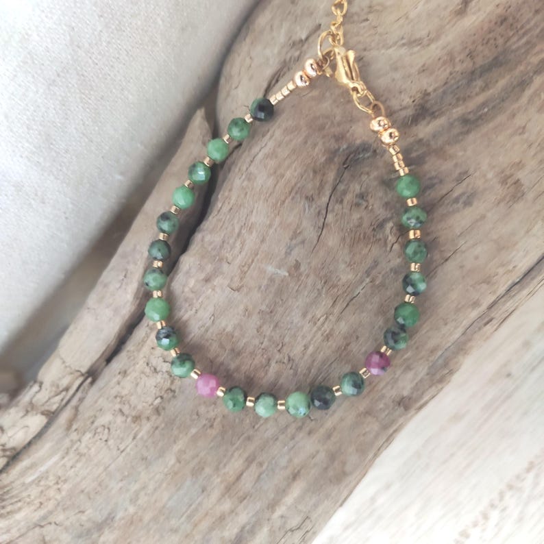 May include: A delicate bracelet featuring green and dark green beads interspersed with small pink beads. The beads are strung on a gold-colored chain with a matching clasp. The bracelet is displayed on a piece of driftwood.