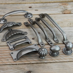 May include: A selection of antique cabinet hardware, including handles, pulls, and knobs. The items have a weathered, grey finish and are displayed on a rustic wooden surface. The hardware is designed in various styles, evoking a vintage aesthetic.