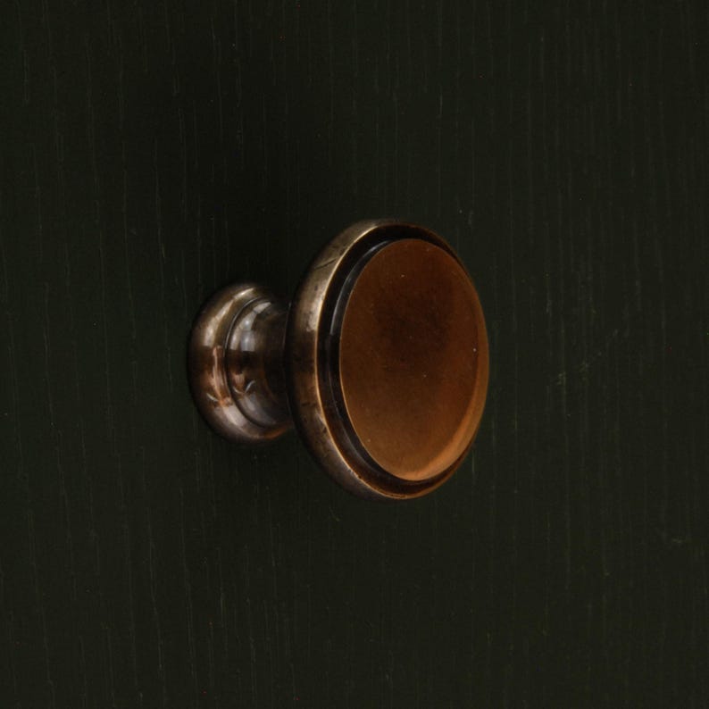 Op de afbeelding: Close-up van een bronzen kastknop met een ovale kern en een cirkelvormige basis. De knop heeft een donkere, antieke afwerking en is geplaatst op een donkergroen oppervlak.