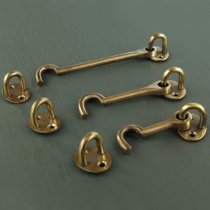 May include: A collection of brass-coloured hook and eye latches and eyelets are displayed on a grey-green surface. The latches feature a curved hook and a rectangular base. The eyelets are circular with a loop and a flat base.