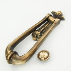 May include: Antique brass door knocker with a decorative floral design. The knocker has a hinged, oval-shaped design with a round, decorative plate.