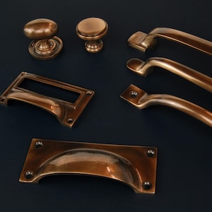 Op de afbeelding: Collectie antieke bronzen kastbeslag, waaronder knoppen, grepen en handgrepen. Het beslag heeft een warme, verouderde afwerking en verschillende ontwerpen, zoals afgeronde knoppen en gebogen grepen. De stukken zijn gerangschikt op een donkerblauwe achtergrond.