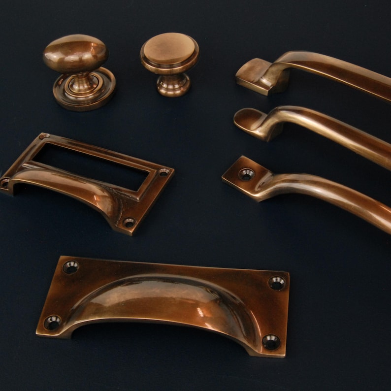 Op de afbeelding: Collectie antieke bronzen kastbeslag, waaronder knoppen, grepen en handgrepen. Het beslag heeft een warme, verouderde afwerking en verschillende ontwerpen, zoals afgeronde knoppen en gebogen grepen. De stukken zijn gerangschikt op een donkerblauwe achtergrond.