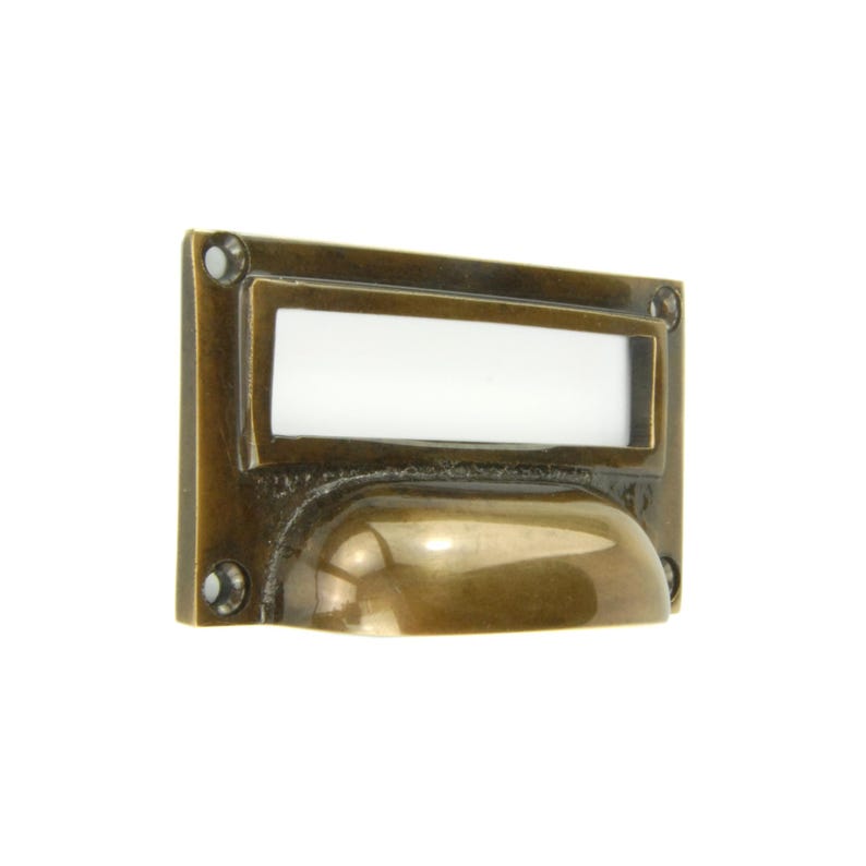 Op de afbeelding: Een rechthoekige messing ladetrekker met een gebogen handvat en een witte labelhouder. De trekker heeft een vintage uitstraling, met zichtbare schroefgaten in elke hoek. Het messing heeft een warme, verouderde patina.