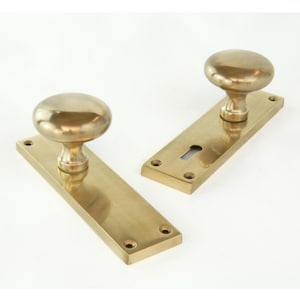May include: Two brass door knobs with rectangular backplates. The knobs are round and the backplates have four screw holes. One backplate has a keyhole. The hardware has a polished finish.