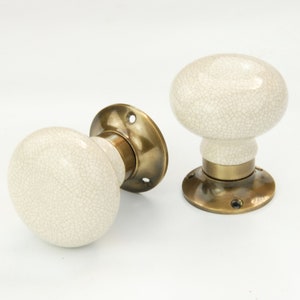 May include: Two white ceramic door knobs with a crackled finish and brass bases. The knobs are round and have a slight indentation in the centre.