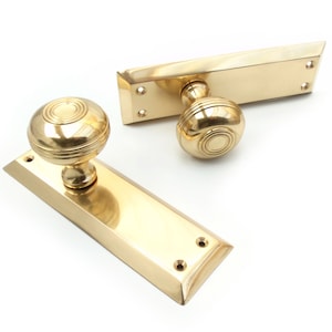 May include: Two polished brass door handles with a round knob design. Each handle has a rectangular backplate with beveled edges and screw holes. The knobs have concentric circle detailing.