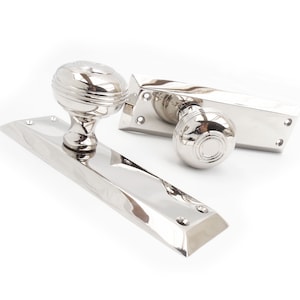 May include: Polished nickel door hardware set. The set includes a door handle with a round knob and a rectangular backplate, a matching door knob, and a rectangular door latch plate. The hardware has a reflective surface.