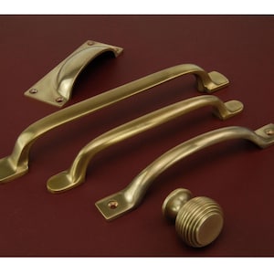 May include: A selection of brass cabinet hardware, featuring curved pull handles, a cup pull, and a round knob. The hardware is in various sizes and shapes, with a warm, golden finish. The items are arranged on a dark red surface.