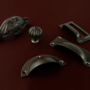 May include: A selection of antique cabinet hardware in various styles. The collection includes a decorative pull with floral detailing, a round knob, and several handle styles, all in a dark, aged metal finish.