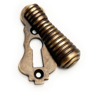 May include: A set of antique brass door hardware, comprising a keyhole plate and a ridged handle. The keyhole plate has a decorative shape and two screw holes. The handle is cylindrical with a spiral design. The hardware is likely for a vintage door.