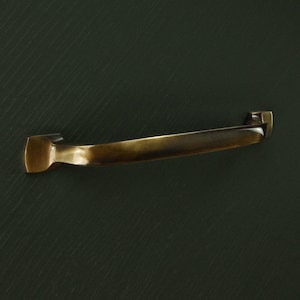 Op de afbeelding: Een gebogen, antiek messing kastgreep met rechthoekige montageplaten. De handgreep heeft een glad, gebogen ontwerp en een warme, goudbruine kleur. De hardware is ontworpen voor meubels.