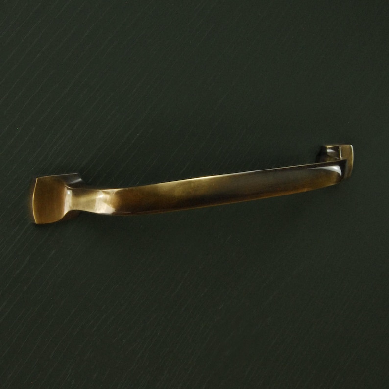 Op de afbeelding: Een gebogen, antiek messing kastgreep met rechthoekige montageplaten. De handgreep heeft een glad, gebogen ontwerp en een warme, goudbruine kleur. De hardware is ontworpen voor meubels.