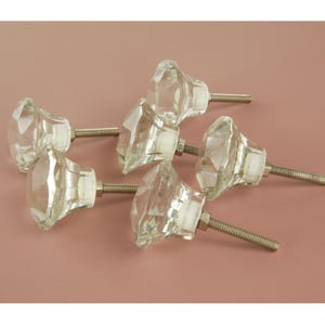 May include: Six clear glass cabinet knobs with faceted diamond-like designs. Each knob has a silver-coloured threaded post and a white base. The knobs are arranged on a light pink background.