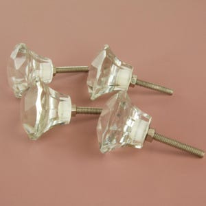May include: Four clear, faceted glass cabinet knobs with silver-coloured metal screws. The knobs have a diamond-like shape and are arranged on a light pink background. These decorative knobs can be used for furniture or other home decor projects.
