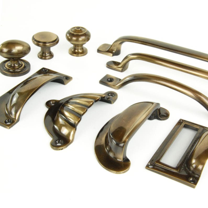 Op de afbeelding: Een verzameling antieke messing kastbeslagen, waaronder knoppen, grepen en handgrepen. De hardware heeft verschillende ontwerpen, van eenvoudige ronde knoppen tot sierlijke, gebogen grepen. De afwerking heeft een warme, verouderde uitstraling.