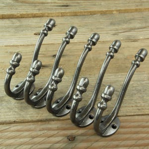 May include: Six antique-style cast iron hooks with acorn finials. The hooks are arranged in a row and are all facing the same direction. The hooks are a dark grey colour and have a rustic finish.