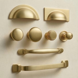 May include: A selection of brass cabinet hardware, including knobs and pulls. The hardware features various designs, such as rounded knobs, a ribbed knob, and cup pulls. The finish is a warm, brushed brass.
