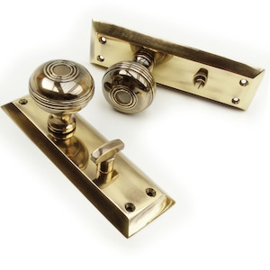 May include: Two antique brass door knobs and plates. The knobs are round with concentric circles, and the plates are rectangular with rounded edges. The hardware is a warm, golden colour, suggesting age and quality. The door hardware is likely for a vintage or classic home.