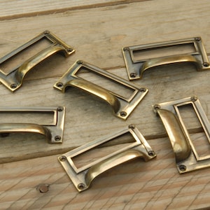 May include: Six antique brass drawer pulls with a rectangular label holder. The pulls have a curved, arched design.