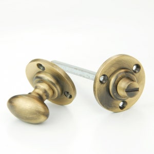 May include: Antique brass door knob set featuring an oval handle and a round backplate. The set includes a connecting rod and a matching backplate with screw detail. The hardware has a vintage aesthetic.