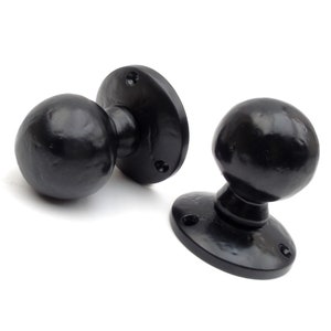 May include: Two black door knobs with round handles and a flat, oval-shaped base. The knobs are made of metal and have a matte finish.