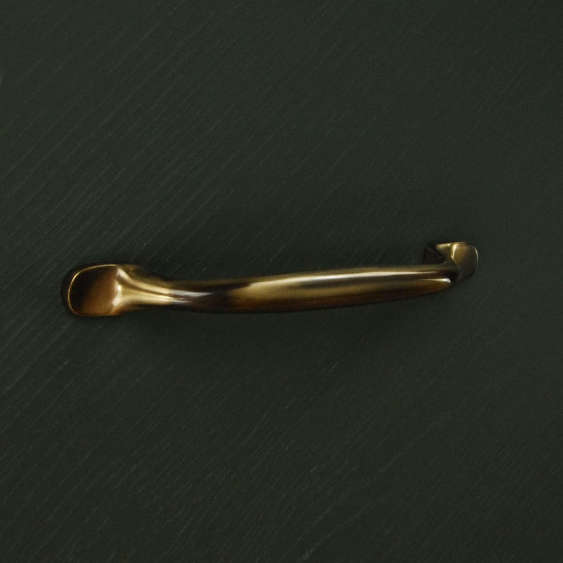 Op de afbeelding: Een bronzen kastgreep met een gebogen ontwerp. De handgreep heeft een gladde, gepolijste afwerking en is geplaatst tegen een donkere, getextureerde achtergrond. Deze greep is geschikt voor laden en kasten.