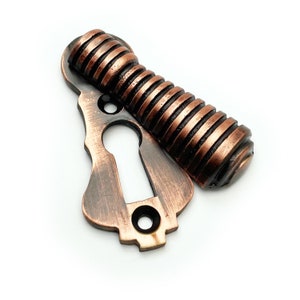 May include: A copper-toned keyhole cover with a decorative, ribbed design. The cover has a keyhole cut-out and a small screw hole.