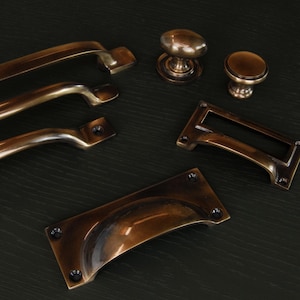 Op de afbeelding: Een assortiment antieke bronzen kastbeslagen, waaronder handgrepen, knoppen en een labelhouder. De hardware heeft een warme, verouderde afwerking en is gerangschikt op een donkere ondergrond. De collectie biedt een verscheidenheid aan stijlen voor meubelrestauratie of nieuwbouw.