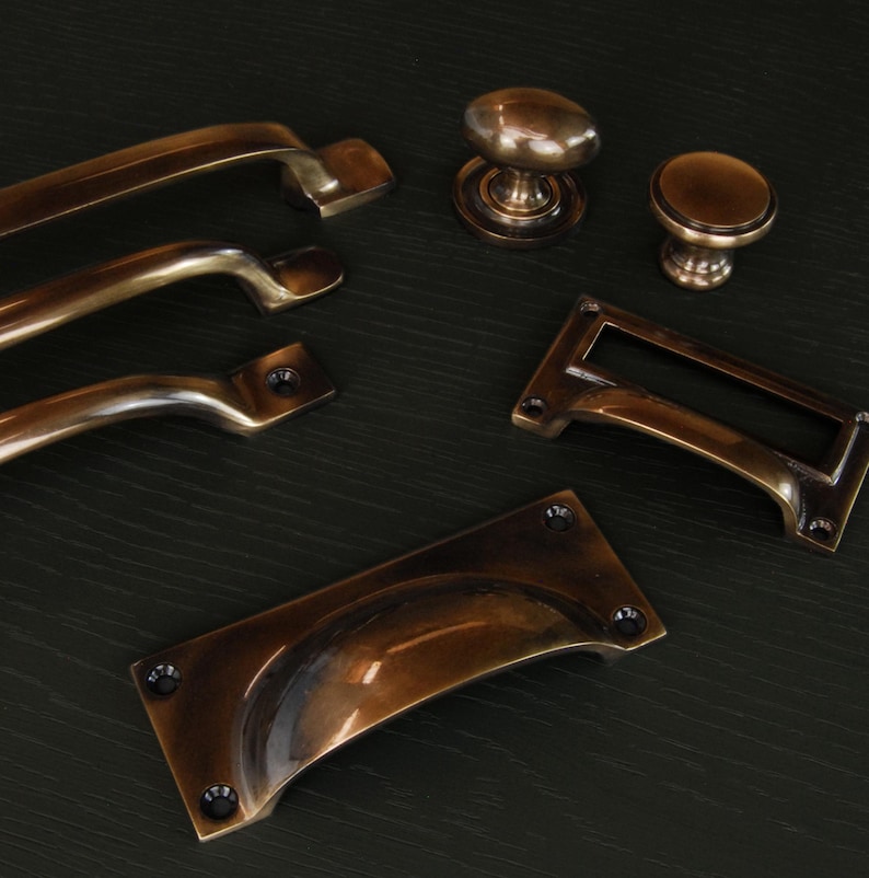 Op de afbeelding: Een assortiment antieke bronzen kastbeslagen, waaronder handgrepen, knoppen en een labelhouder. De hardware heeft een warme, verouderde afwerking en is gerangschikt op een donkere ondergrond. De collectie biedt een verscheidenheid aan stijlen voor meubelrestauratie of nieuwbouw.