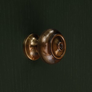 Op de afbeelding: Close-up van een messing deurknop tegen een donkergroene achtergrond. De deurknop heeft een afgeronde vorm met een cirkelvormig ontwerp aan de voorkant. Het metaal heeft een warme, antieke afwerking.