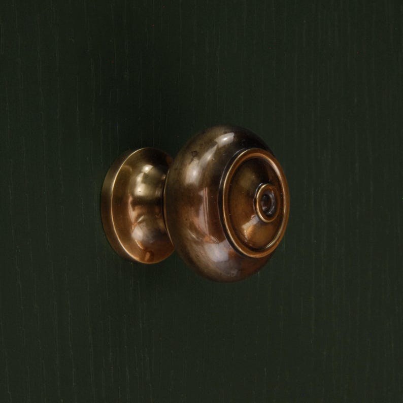 Op de afbeelding: Close-up van een messing deurknop tegen een donkergroene achtergrond. De deurknop heeft een afgeronde vorm met een cirkelvormig ontwerp aan de voorkant. Het metaal heeft een warme, antieke afwerking.