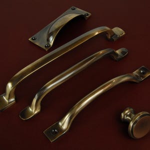 Op de afbeelding: Een assortiment antieke messing kastbeslagen, waaronder verschillende gebogen handgrepen, een rechthoekige achterplaat en een ronde knop. De hardware heeft een warme, verouderde afwerking en is geplaatst op een donkerroodbruin oppervlak.