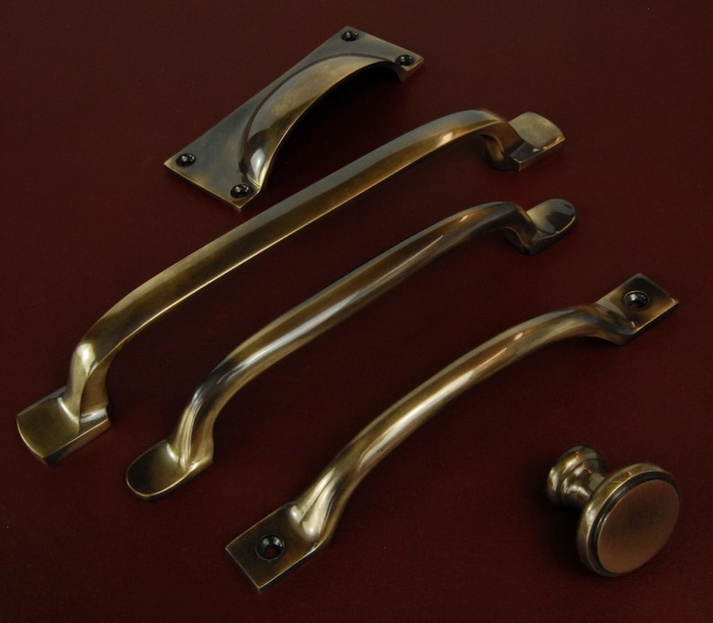 Op de afbeelding: Een assortiment antieke messing kastbeslagen, waaronder verschillende gebogen handgrepen, een rechthoekige achterplaat en een ronde knop. De hardware heeft een warme, verouderde afwerking en is geplaatst op een donkerroodbruin oppervlak.