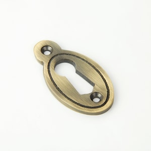 May include: An antique brass keyhole cover. The oval-shaped cover features a keyhole in the centre and two small mounting holes. The metal has a distressed finish. The dimensions are approximately 7 cm in length.