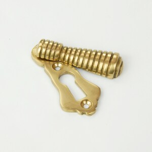 May include: A brass keyhole cover with a decorative, ornate design. The cover has a separate, ribbed knob that can be used to open the keyhole.