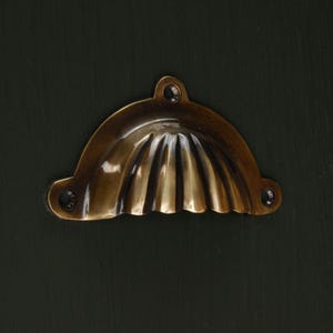 Op de afbeelding: Een messingkleurige, halvemaanvormige trekker met een geribbeld ontwerp. De hardware heeft drie schroefgaten voor montage en is geplaatst tegen een donkergroene achtergrond. Deze decoratieve trekker is geschikt voor kasten en laden.