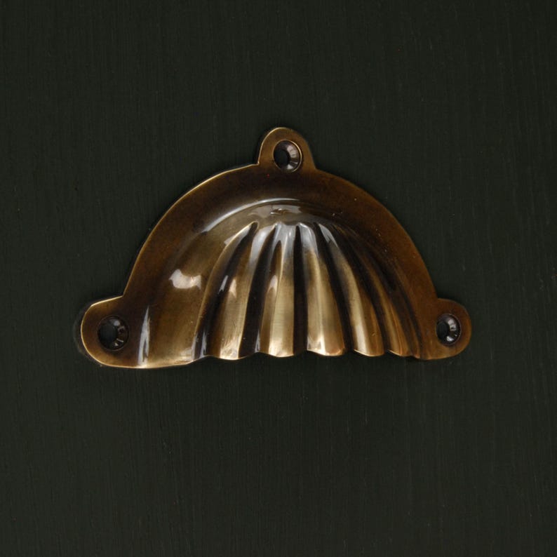 Op de afbeelding: Een messingkleurige, halvemaanvormige trekker met een geribbeld ontwerp. De hardware heeft drie schroefgaten voor montage en is geplaatst tegen een donkergroene achtergrond. Deze decoratieve trekker is geschikt voor kasten en laden.