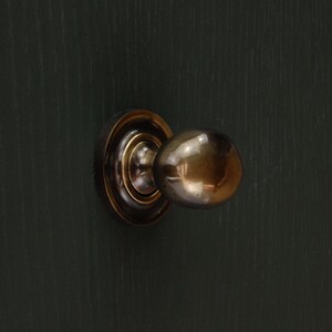 Op de afbeelding: Een messing deurknop met een ronde handgreep en een cirkelvormige achterplaat, gemonteerd op een donkergroene houten deur. Het deurbeslag heeft een vintage uitstraling. De knop heeft een diameter van ongeveer 5 cm.
