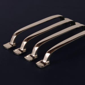 May include: Four polished chrome cabinet pulls. Each pull has a rectangular base and a curved handle. The shiny metal reflects light, creating highlights and shadows. These cabinet pulls are designed for furniture or cabinetry.